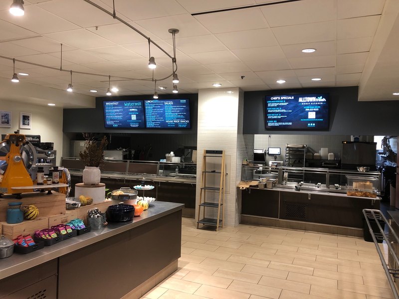 Hero-style establishing shot of the whole cafe: three glowing digital menu boards above the serving lines, track lighting, and the merchandised front counter.