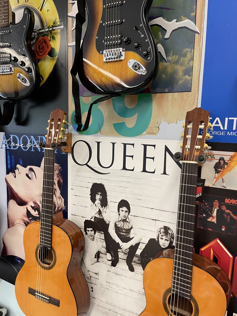 Colorful detail of sunburst electric guitars and classical acoustics on wall hangers over Queen, Madonna, and AC/DC posters.