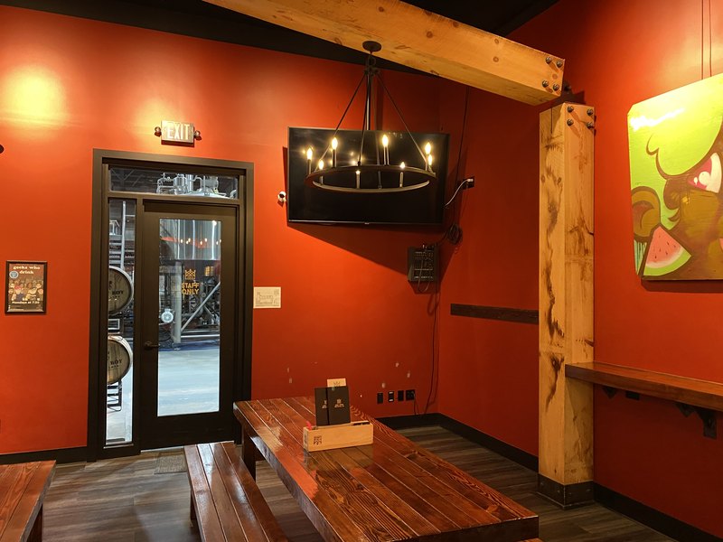 Corner detail of a wall-mounted flat-panel TV with a compact audio mixer mounted on the wall beside it and a ring chandelier in front, with the barrel room visible through the interior window.