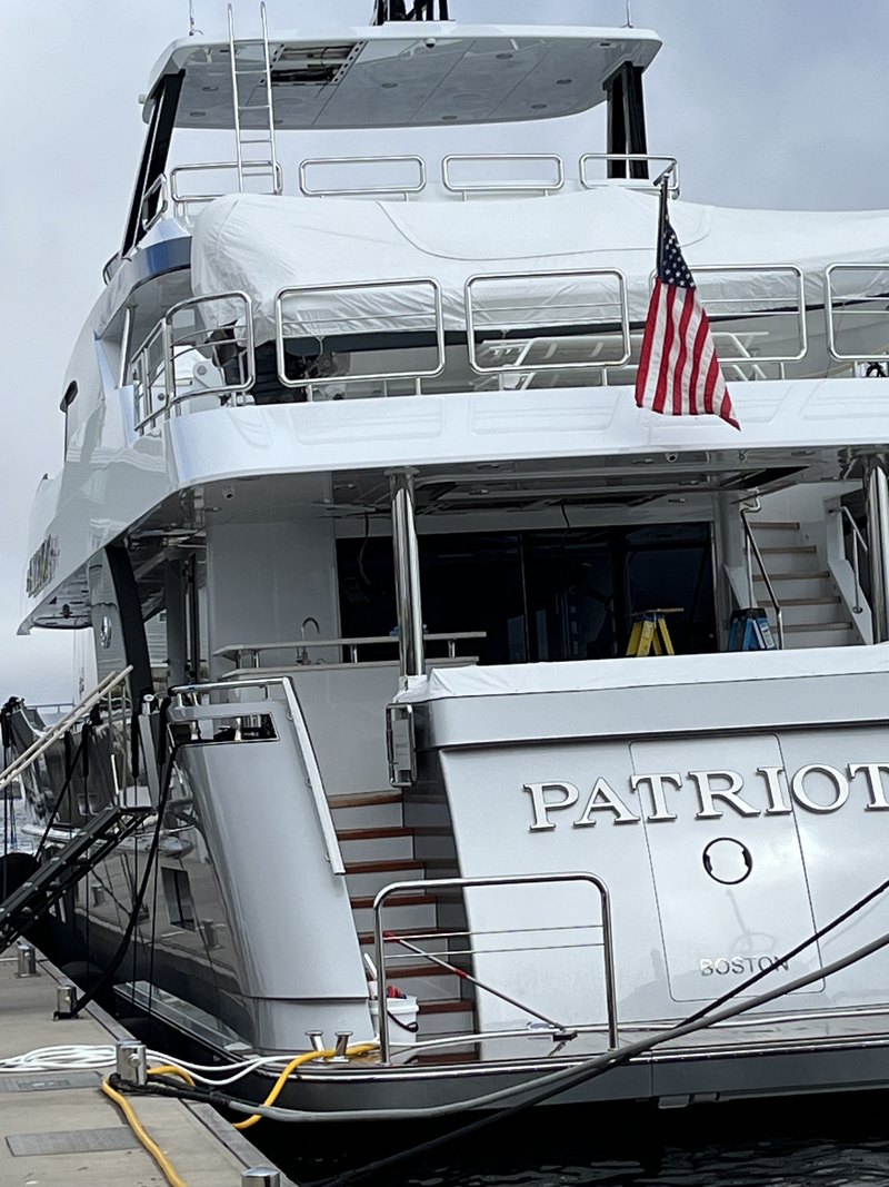 Stern view of the yacht with 'PATRIOT — BOSTON' transom lettering, American flag, tender on the boat deck, and shore-power cables from the install still connected.
