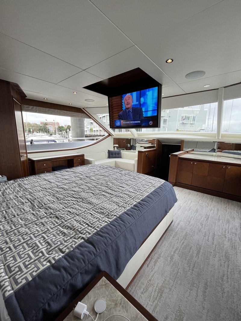 Master stateroom with the wall-mounted TV playing live TV, in-ceiling speaker and downlights above, and panoramic harbor-view windows behind the bed.