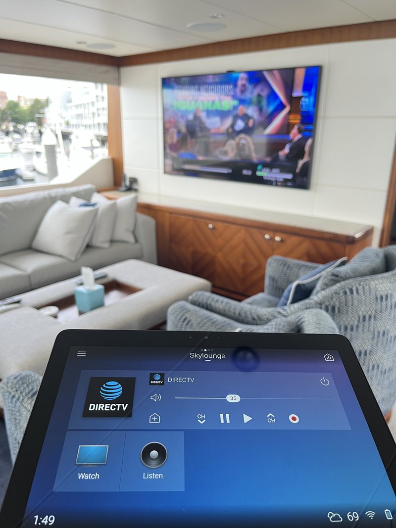 Close-up of a touchscreen control panel on the 'Skylounge' zone showing DIRECTV watch/listen controls and volume, with the TV playing in the background.