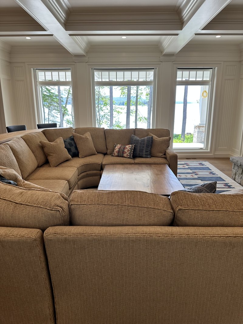 Same great room angle with the motorized shades raised, daylight and lake/tree views pouring through the tall windows above the sectional — the 'after' half of the shades-down/shades-up pair; image is stored rotated 90 degrees sideways and needs rotation before use.